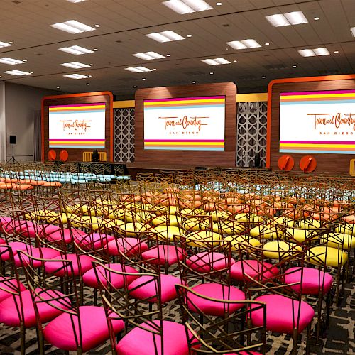 A colorful conference room setup with rows of chairs featuring pink, orange, and yellow cushions, facing three large screens displaying text.