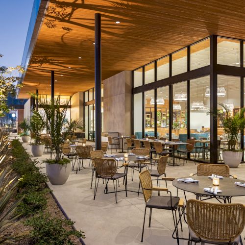 An elegant restaurant with outdoor seating, modern furniture, large windows, and lush plants, set in a serene evening atmosphere.