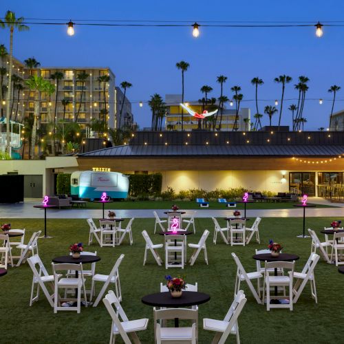 An outdoor venue with white chairs and tables, neon flamingo lights, surrounded by palm trees and string lights at dusk.