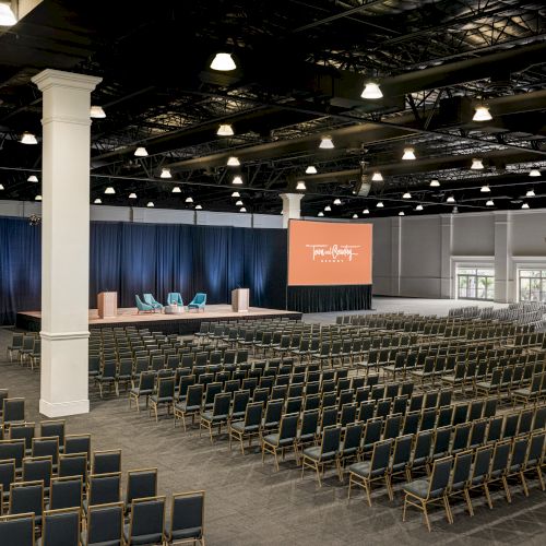 An empty conference hall with rows of chairs, a stage with chairs, and large screens displaying the text "Think Better, Live Better."