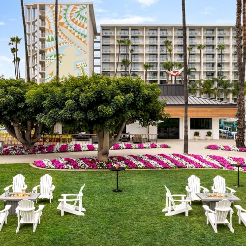 The image shows a garden with white chairs, a large tree, flower beds, and a building in the background.