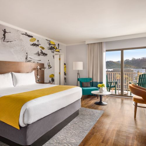 The image shows a hotel room with a bed, colorful wall art, chairs, a table, a TV, and a balcony with a scenic view outside.