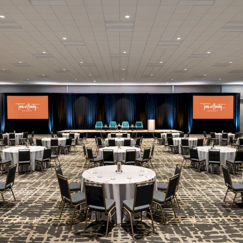 The image shows a conference room set up with round tables and chairs, a stage, and two projectors displaying orange screens.