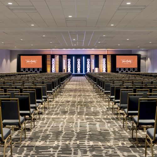 A large conference room filled with rows of empty chairs facing a stage with screens displaying text on both sides.