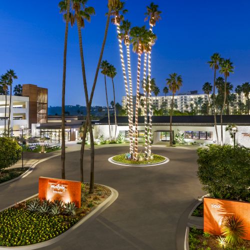 The image shows the entrance of a contemporary hotel with palm trees, landscaped gardens, and modern art sculptures in an urban setting.