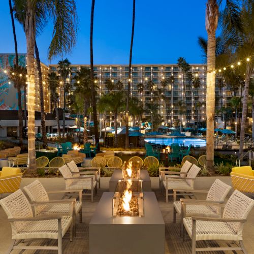 An outdoor lounge area with a fire pit, surrounded by yellow and white chairs, with palm trees, string lights, and a hotel building in the background.