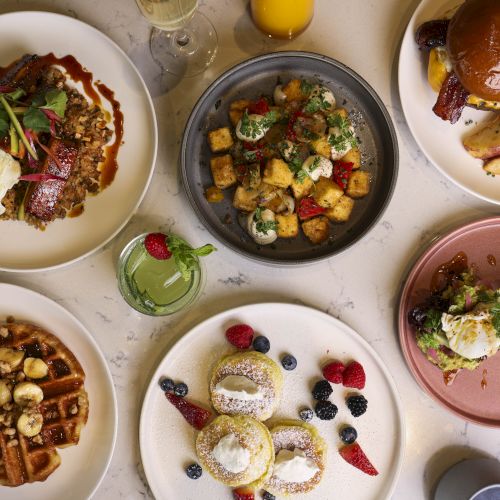 A table with a variety of dishes including waffles, eggs Benedict, a salad, roasted potatoes, a burger, stuffed pastries, and a bowl of croutons and vegetables.