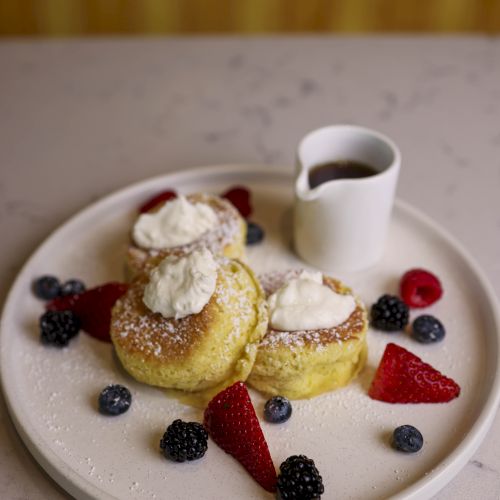 A plate with three pancakes topped with whipped cream, surrounded by strawberries, raspberries, blueberries, and blackberries, with a small jug of syrup.