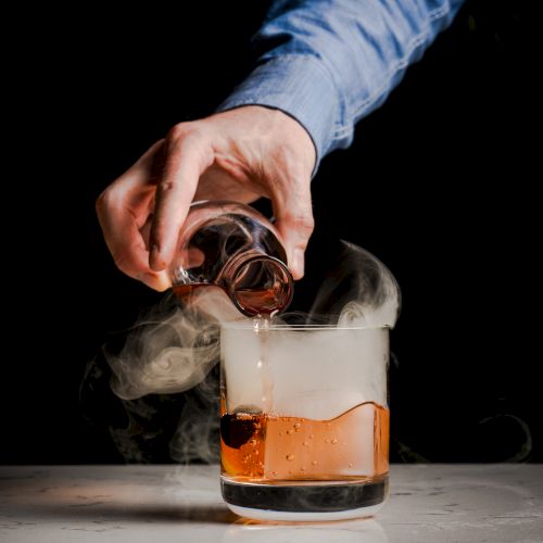 A hand in a blue shirt is pouring liquid from a small glass into a larger glass with smoke rising from it, set on a dark background.