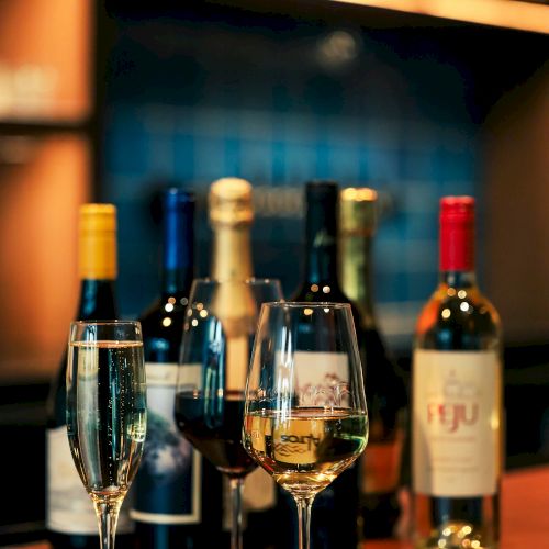 The image shows several wine bottles and three filled wine glasses on a wooden counter, likely in a bar or restaurant setting.