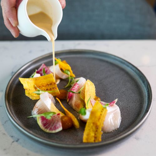 A hand is pouring a creamy sauce from a small pitcher onto a plate of beautifully arranged food with colorful garnishes and slices.