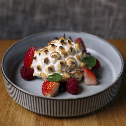 A plate with a dessert covered in torched meringue, garnished with strawberries, raspberries, and mint leaves, sits on a wooden table.