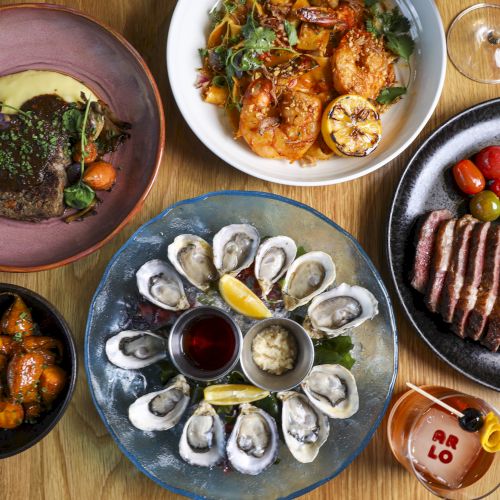 The image shows a table with various dishes: steak, oysters, shrimp pasta, roasted vegetables, cherry tomatoes, a glass of wine, and a cocktail.