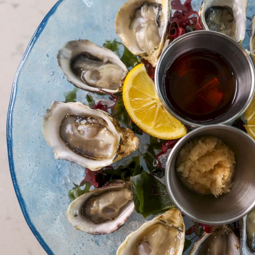 A plate of fresh oysters with lemon, sauce, and garnishes, presented on a blue dish, arranged around two condiment containers.
