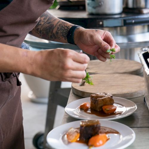A chef is garnishing two plated dishes with fresh herbs, likely finishing touches for a gourmet meal in a professional kitchen environment.
