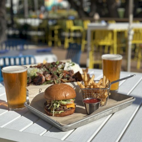 The image shows a delicious meal with a burger, fries, and two glasses of beer on a white table, set in an outdoor dining area.
