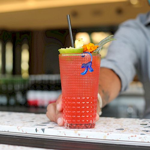 A person wearing a mask is holding a colorful, garnished drink in a decorative glass with a straw at a bar or cafe setting.