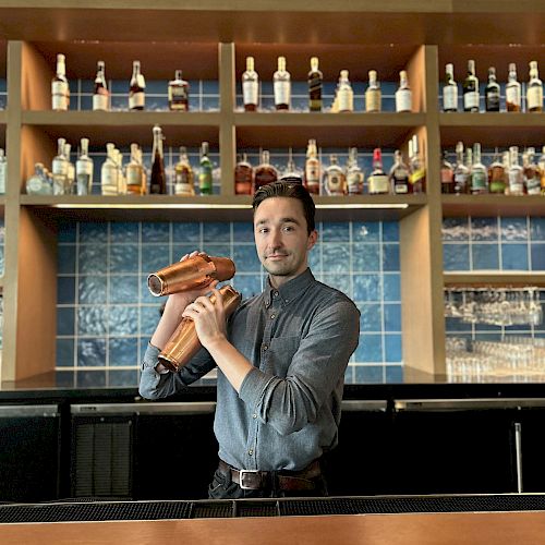 A bartender stands behind a bar with shelves of liquor bottles, holding a cocktail shaker, ready to prepare a drink.