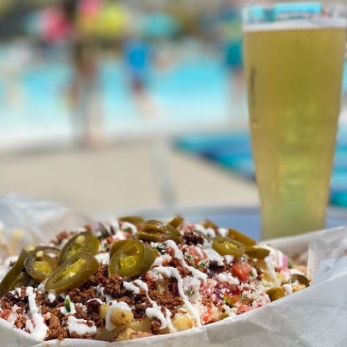 A delicious-looking tray of nachos topped with jalapeños, veggies, and sauce sits next to a tall glass of beer by a poolside.