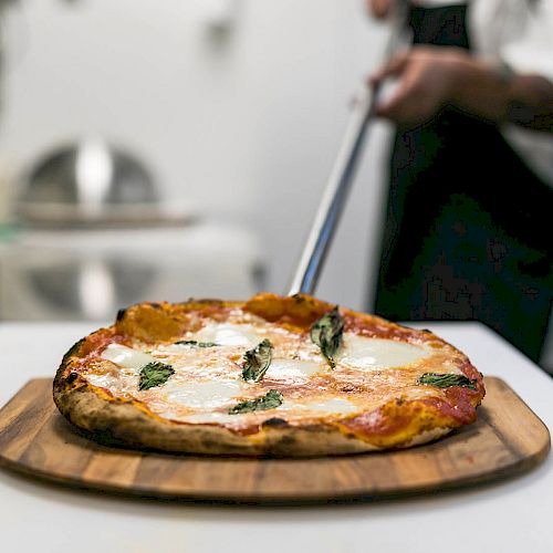 A freshly baked pizza with cheese and basil on a wooden board is being served in a kitchen, with a person in the background.