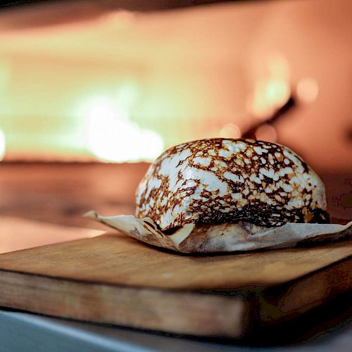A baked, brown-topped pastry rests on a wooden board, with a fire burning in the background.