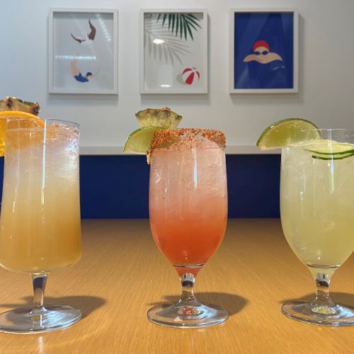 Three cocktails are lined up on a wooden table, garnished with fruit slices. Artwork featuring beach scenes is displayed on the wall behind them.