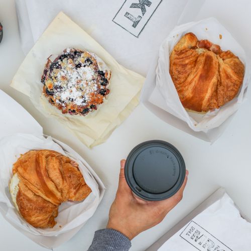 The image shows three croissants and a pastry with powdered sugar and berries, along with a hand holding a coffee cup, on a white table.