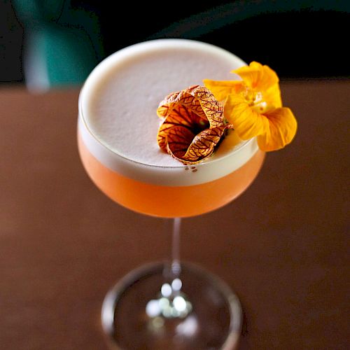 A cocktail glass with a frothy orange drink, garnished with a dried citrus slice and yellow flowers, sits on a brown surface.
