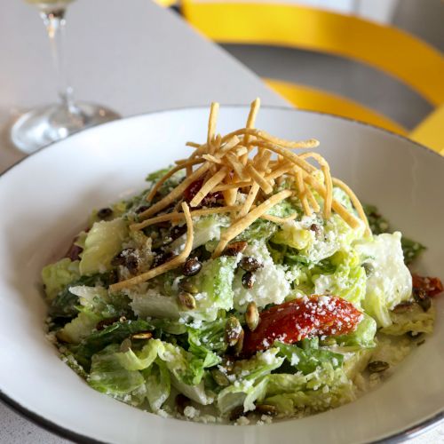 The image shows a fresh salad garnished with strips of fried tortilla served in a white bowl, accompanied by a glass of white wine on a table.