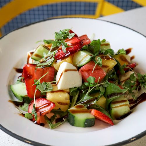 A fresh salad with mixed greens, cucumber, strawberries, mozzarella, and a drizzle of balsamic dressing, served in a white bowl on a table.