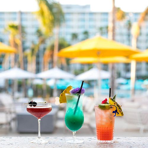 Three colorful cocktails sit on a bar with a tropical resort background, featuring bright yellow umbrellas and palm trees in the distance.