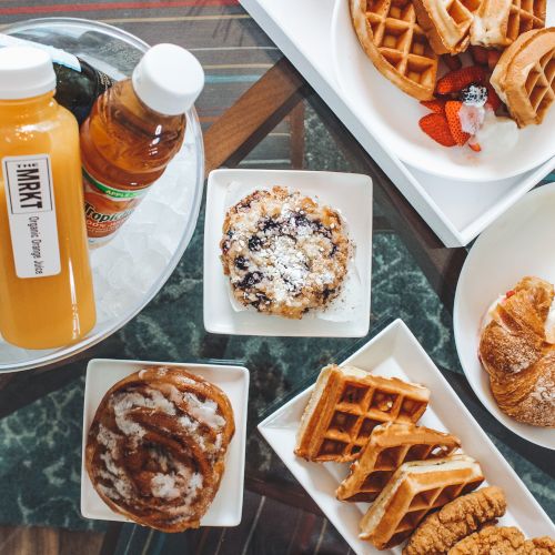 The image shows a breakfast spread with waffles, croissant, fruit, pastries, a muffin, orange juice bottles, and a small bottle of syrup.