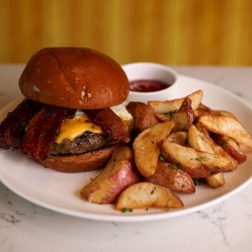 A cheeseburger with bacon lies next to seasoned potato wedges on a white plate, accompanied by a small container of dipping sauce.