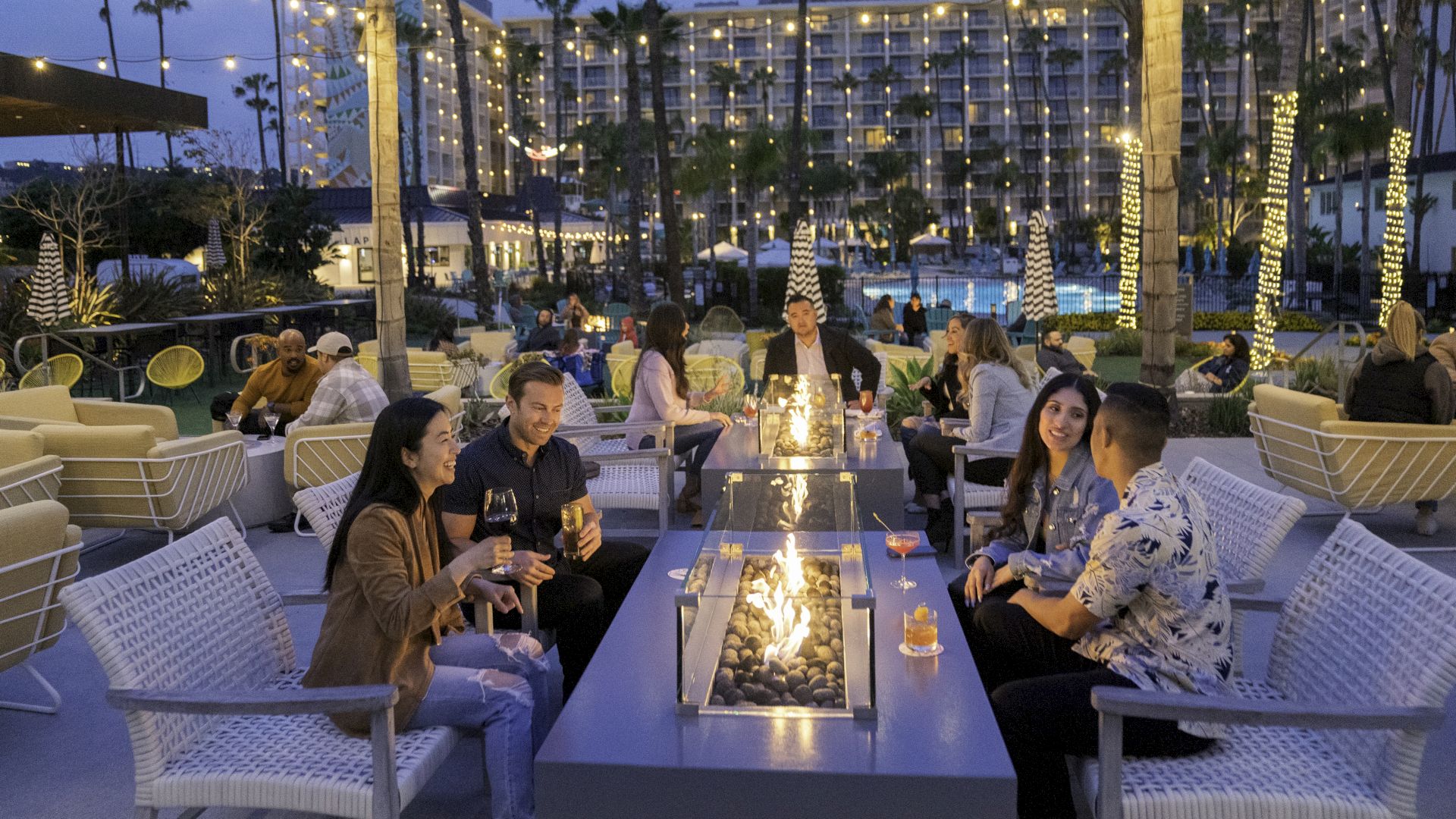 People are socializing around a fire pit in an outdoor lounge area, with a lit pool and a multi-story building in the background.