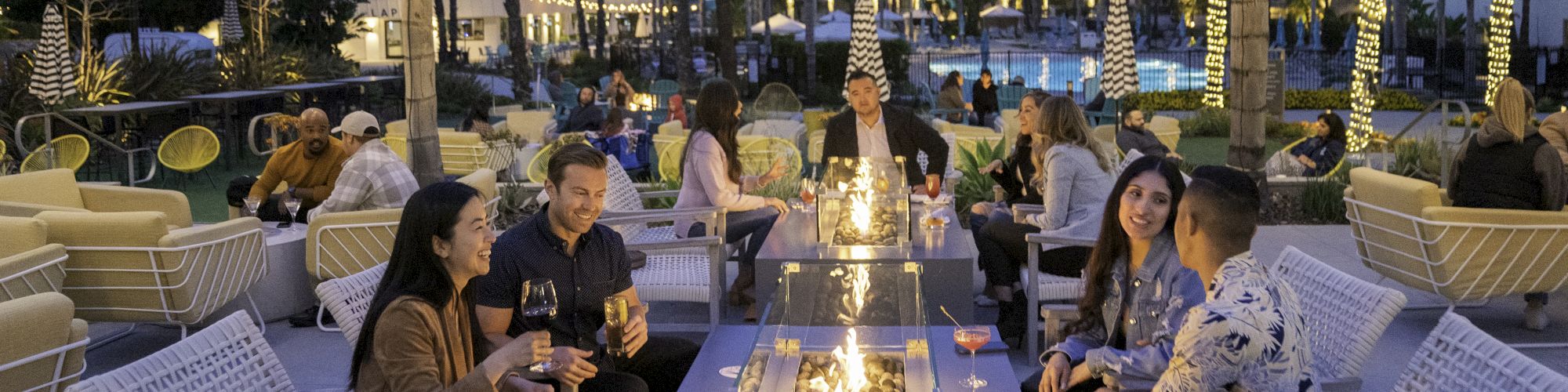 People are socializing around a fire pit in an outdoor lounge area, with a lit pool and a multi-story building in the background.