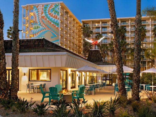 An outdoor seating area with teal chairs and tables outside a building, with tall palm trees and a multi-story hotel with a colorful mural in the background.