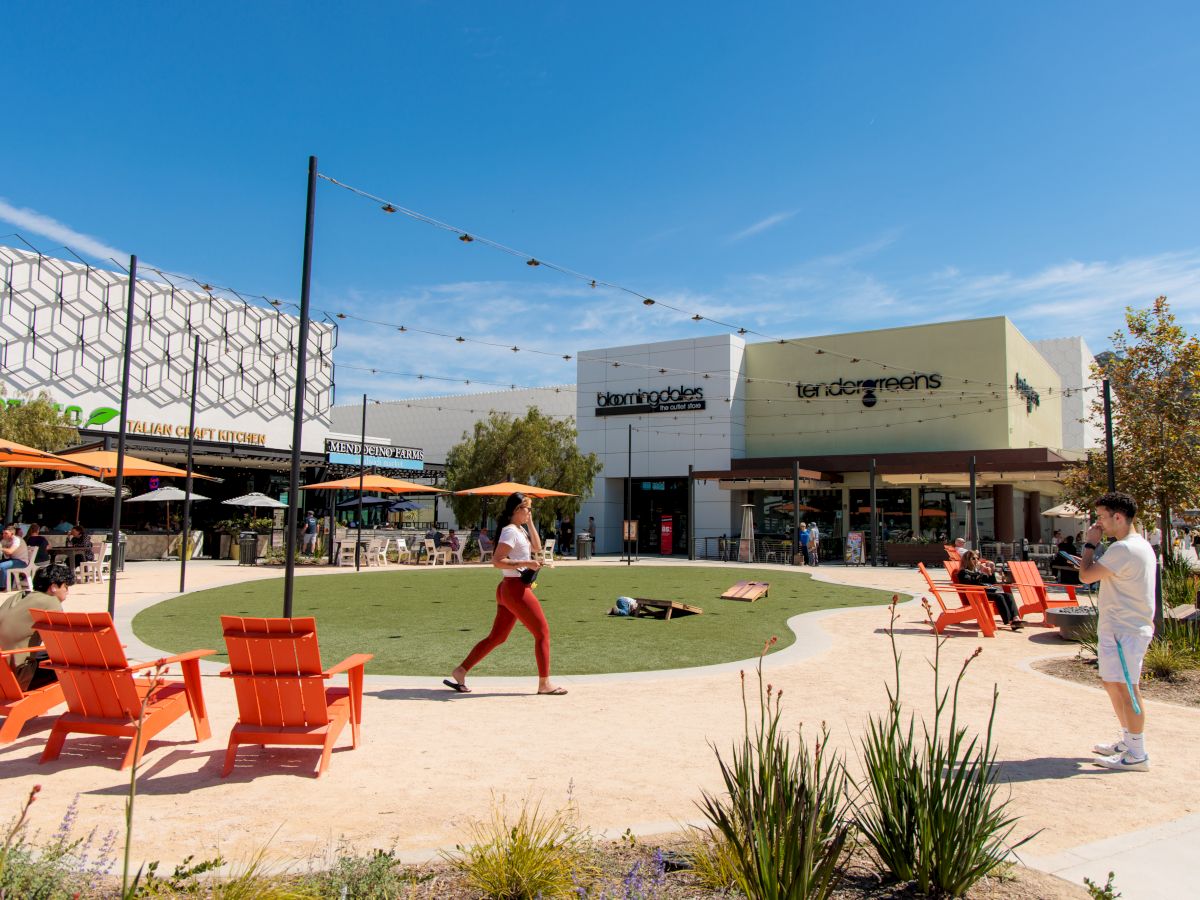 Outdoor shopping plaza with orange deck chairs, people walking, and storefronts labeled Bloomingdale&rsquo;s and Tender Greens under a blue sky.