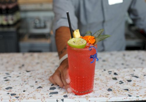 A person is holding a red cocktail garnished with lime, pineapple, and an orange flower, with a straw and a blue stirrer on a speckled countertop.