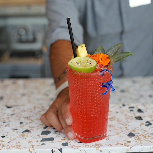 A person is holding a red cocktail garnished with lime, pineapple, and an orange flower, with a straw and a blue stirrer on a speckled countertop.