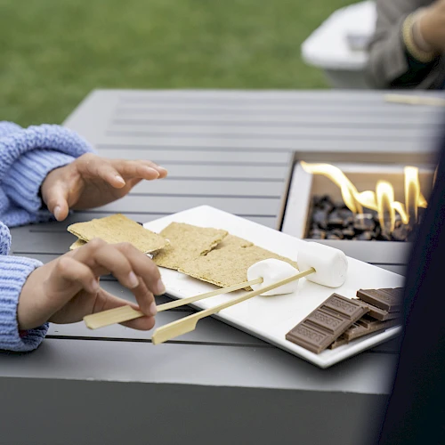 Two people are making s'mores at an outdoor fire pit, with graham crackers, marshmallows, and chocolate on a plate.