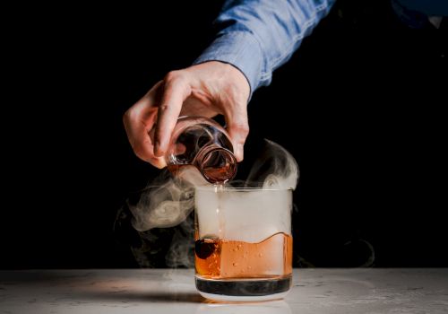 A hand pouring a dark liquid from a small glass into a larger glass emitting smoke, creating a visually intriguing effect.