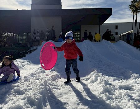 Kids play in a snowy courtyard, rolling on sledding tubes, with a modern building and sunny sky in the background.