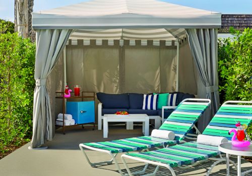 A poolside cabana with striped lounge chairs, a small table with a pink flamingo float, and a shaded area with a sofa and storage cabinet.
