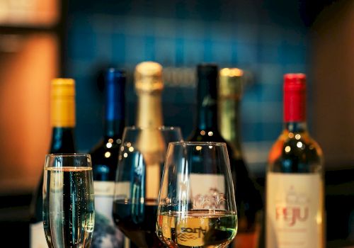 The image shows a bar setup with various bottles of wine and three filled wine glasses placed on a wooden counter.