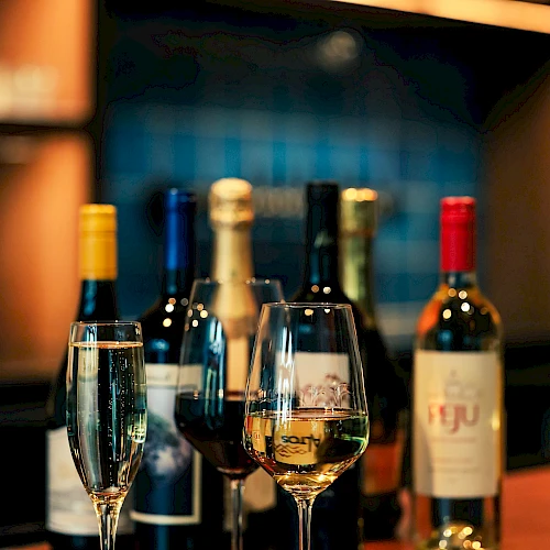 The image shows a bar setup with various bottles of wine and three filled wine glasses placed on a wooden counter.