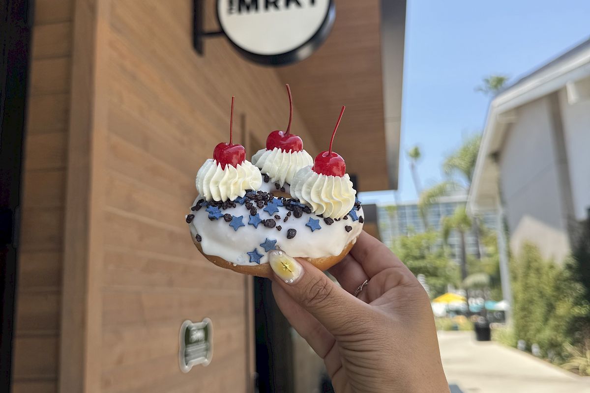A hand holds a decorated donut with cherries and blue stars outside an EMRKT storefront.