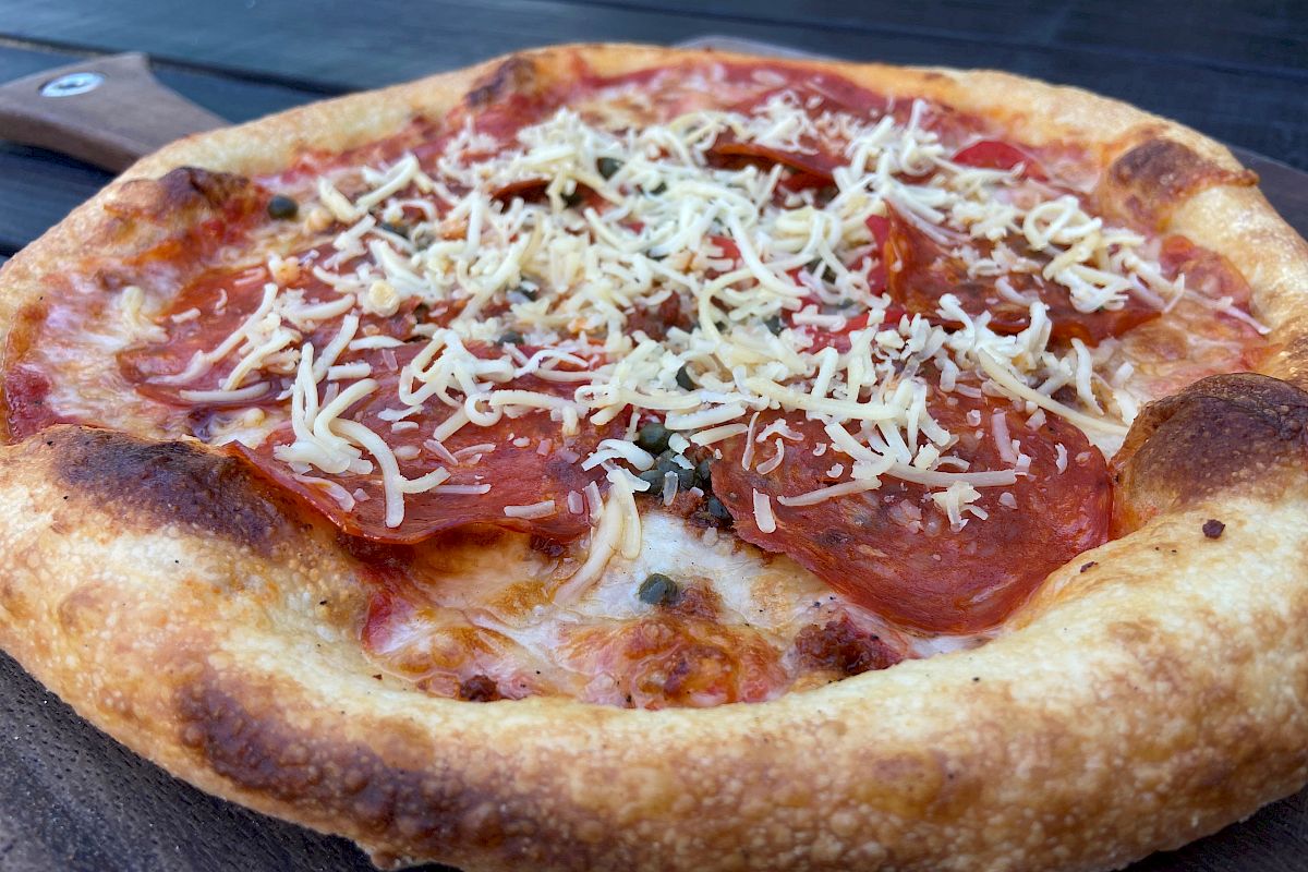 A wood-fired pizza with tomato sauce, mozzarella, peppers, olives, and herbs on a metal tray.