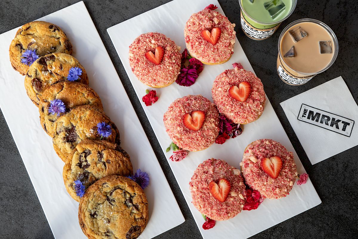 A platter of chocolate-chip cookies, strawberry-dusted donuts with strawberry halves, and two iced drinks on the table.