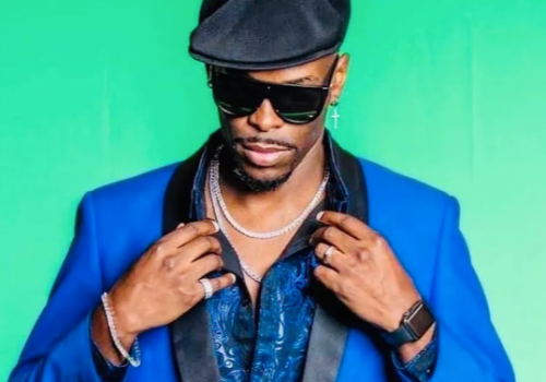 A person wearing a blue blazer, black shirt, black hat, and sunglasses stands against a green background, adjusting their collar with both hands.