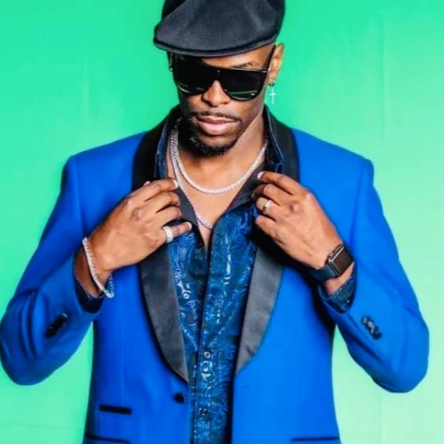 A person wearing a blue blazer, black shirt, black hat, and sunglasses stands against a green background, adjusting their collar with both hands.